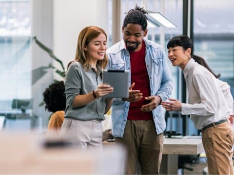 3 colleagues are looking at a tablet while standing up next to a desk in an open space office