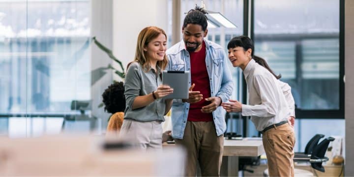 3 colleagues are looking at a tablet while standing up next to a desk in an open space office