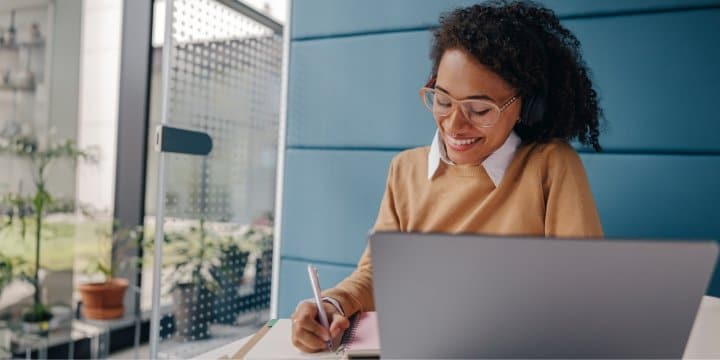 A woman with headphones writing down in her notebook while listening to a virtual meeting in her cubicle